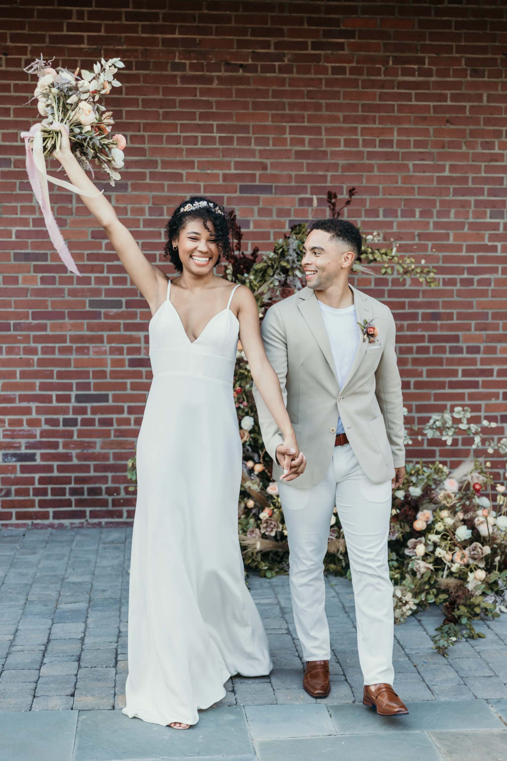 wedding photographer, a delighted bride holds groom's hand, her other hand holds a floral bouquet in the air in celebration